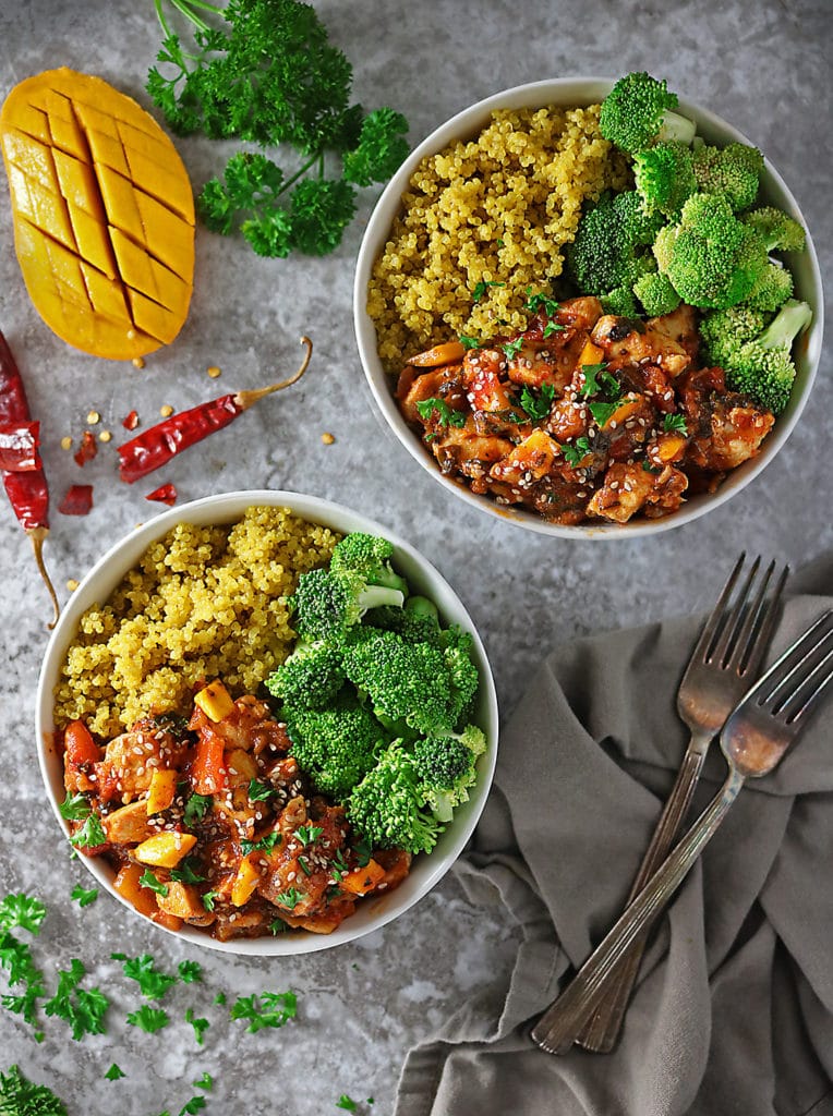 Overhead photo of 2 bowls with mango chicken, turmeric quinoa and broccoli with cut mango and parsley and chili flake garnish