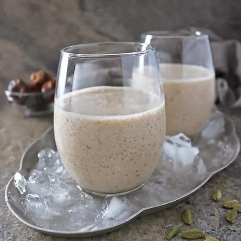 Photo of 2 glasses with date shake in them and cardamom pods scattered in front