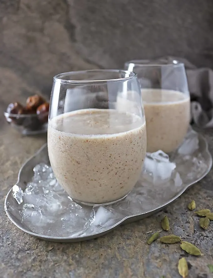 Photo of 2 glasses with date shake in them and cardamom pods scattered in front