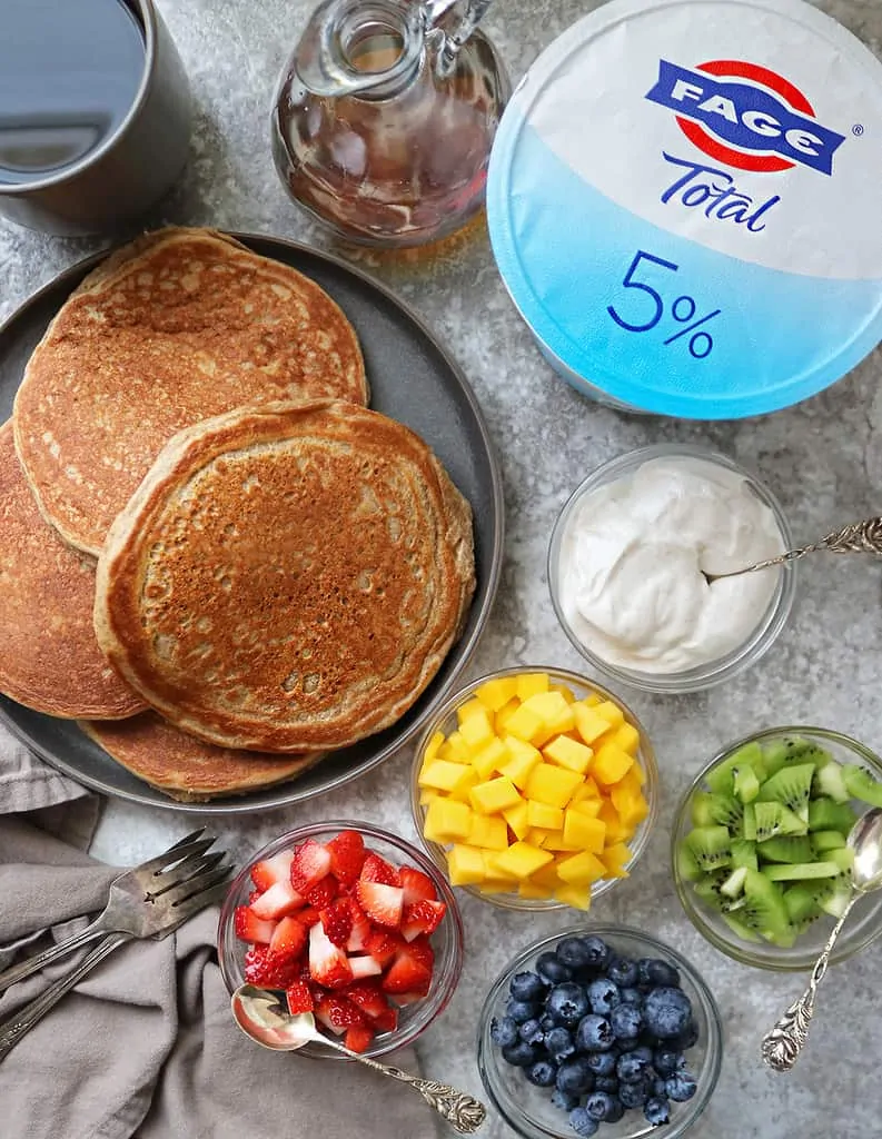 Tasty gluten free yogurt pancakes with bowls of fruit and fage laid out on a table.