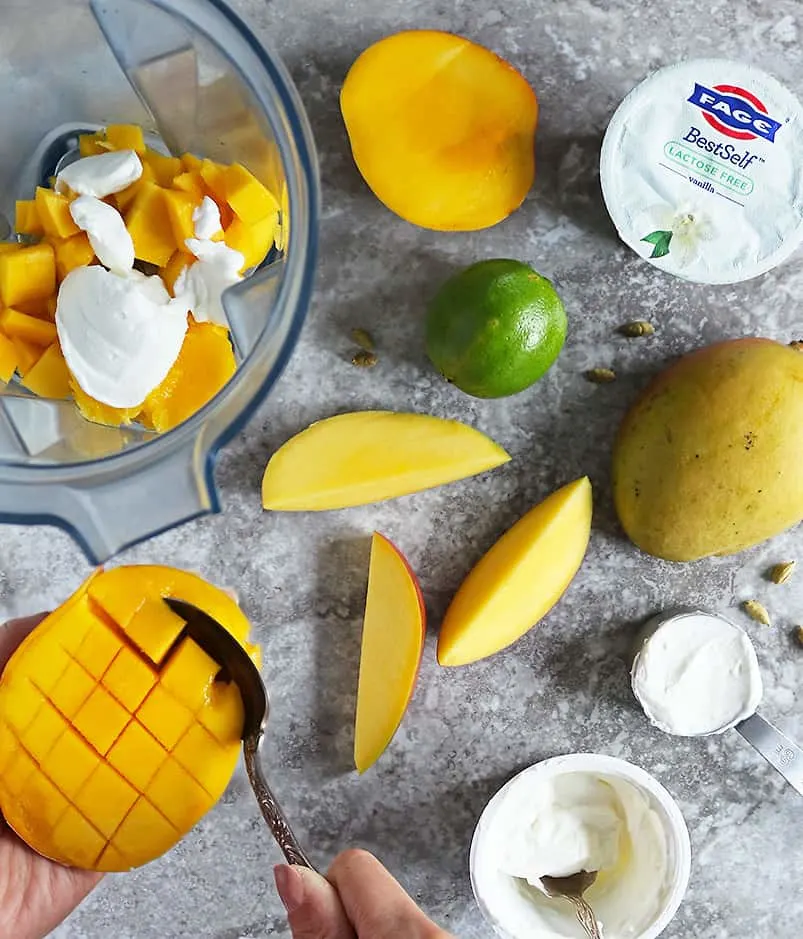 scooping out mango to place in the blender while being surrounded by the ingredients to make mango lassi.