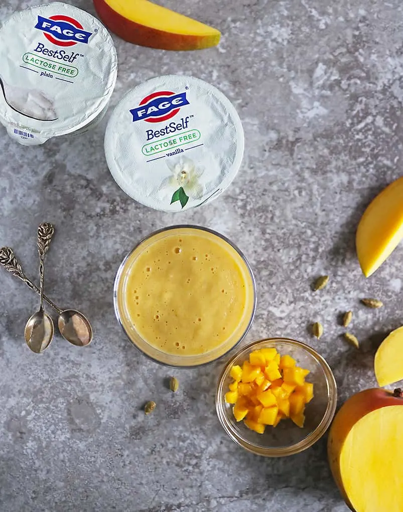 overhead photo of 2 containers of FAGE along with a glass of mango lassi and a bowl with chunks of mango