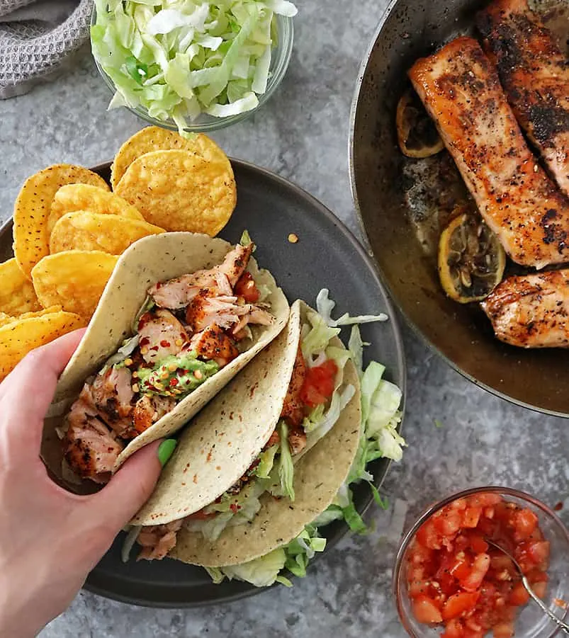 Hand reaching to grab a taco from a plate of two tacos with chips, salmon, lettuce, tomatoes, and gauc on the side.