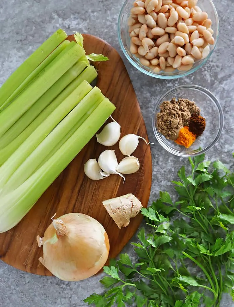 10 ingredients needed to make celery soup laid out on a cutting board on a grey background.