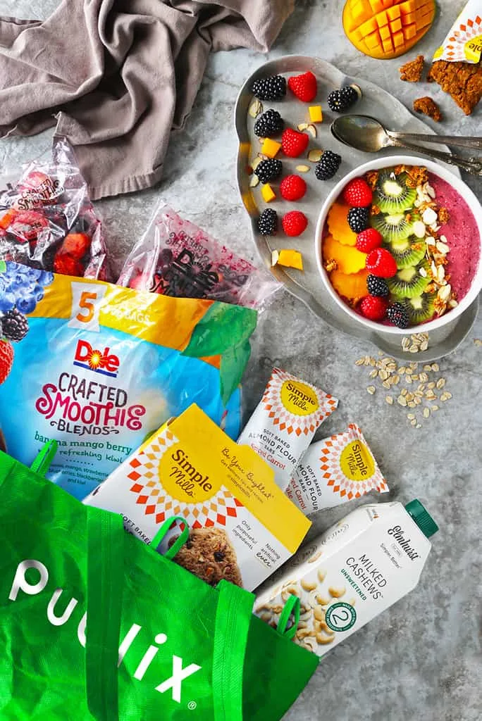 An oatmeal smoothie bowl is in a bowl and it is topped with colorful fruits. Next to it is a publix bag with ingredients used to make this oatmeal smoothie bowl.