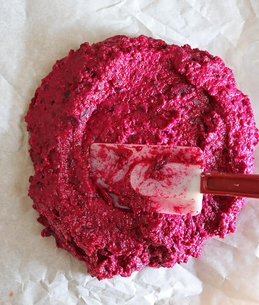 Shaping beet crust pizza dough with a spatula on a parchment lined baking tray.