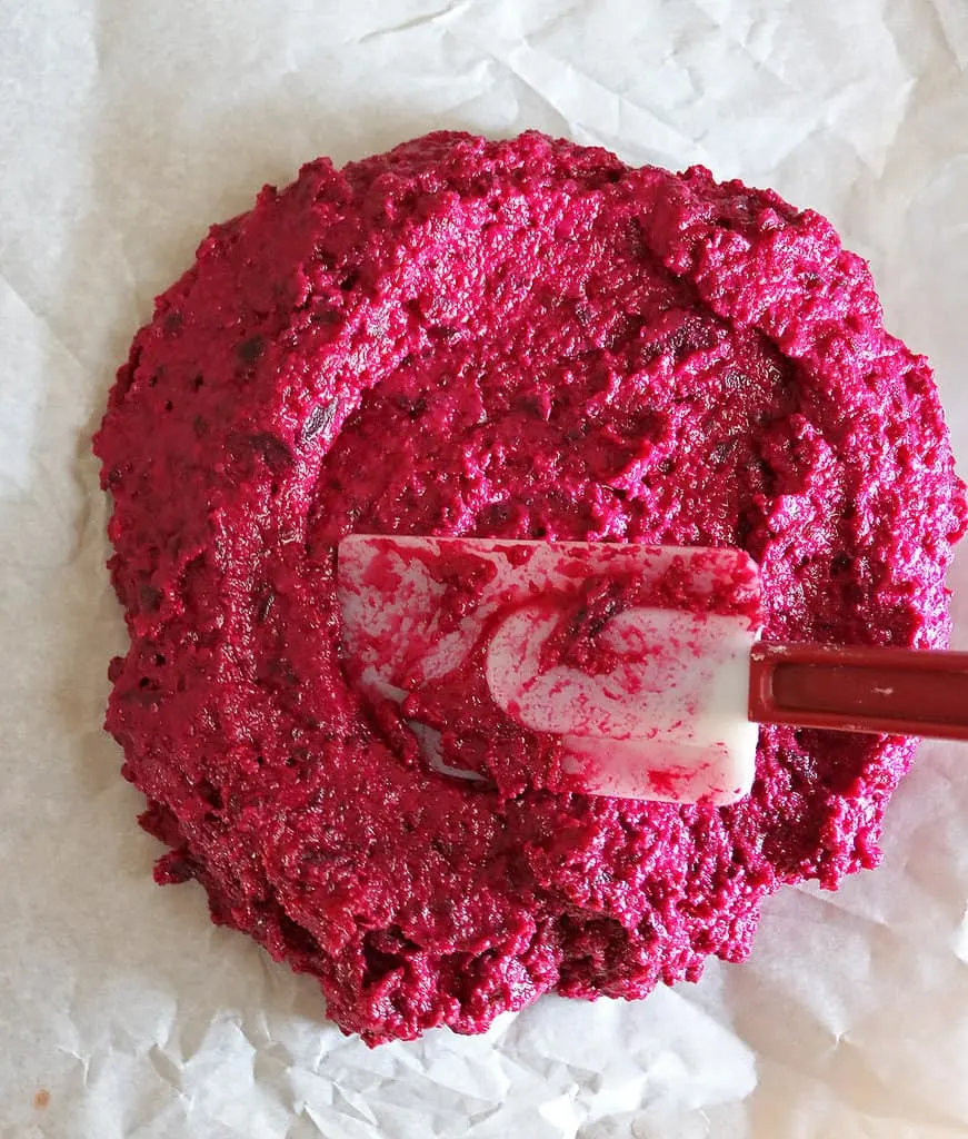 Shaping beet crust pizza dough with a spatula on a parchment lined baking tray.
