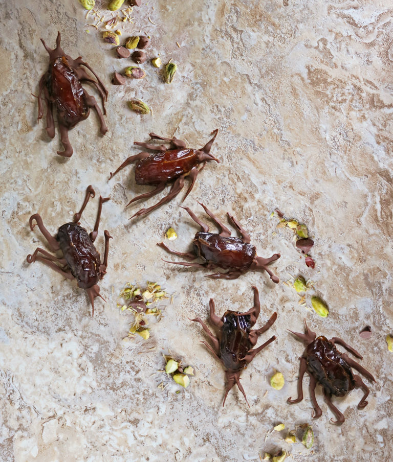 An overhead view of a brown marble board with 6 Dubai Chocolate Cockroaches with pistachios and toasted kataifi tossed around.