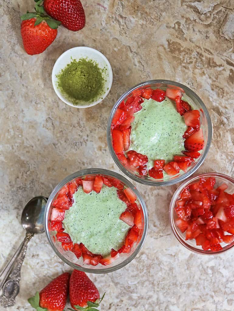 2 servings of Delicious creamy matcha chia pudding with yogurt and fresh strawberries on a brown background with a small bowl with matcha powder near these.