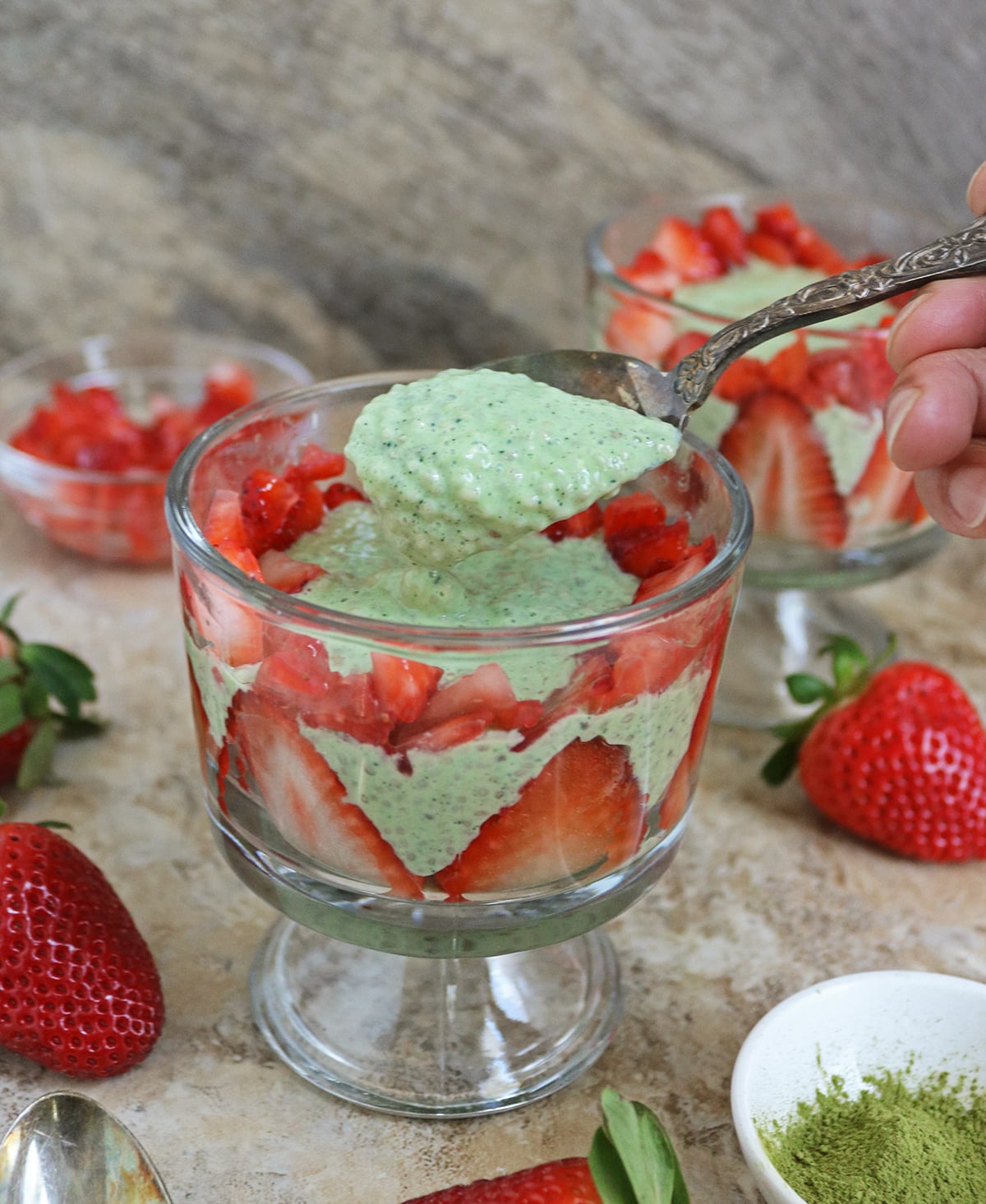 Taking a spoonful of the best matcha chia pudding with yogurt and fresh strawberries from a glass bowl against a marble brown background.