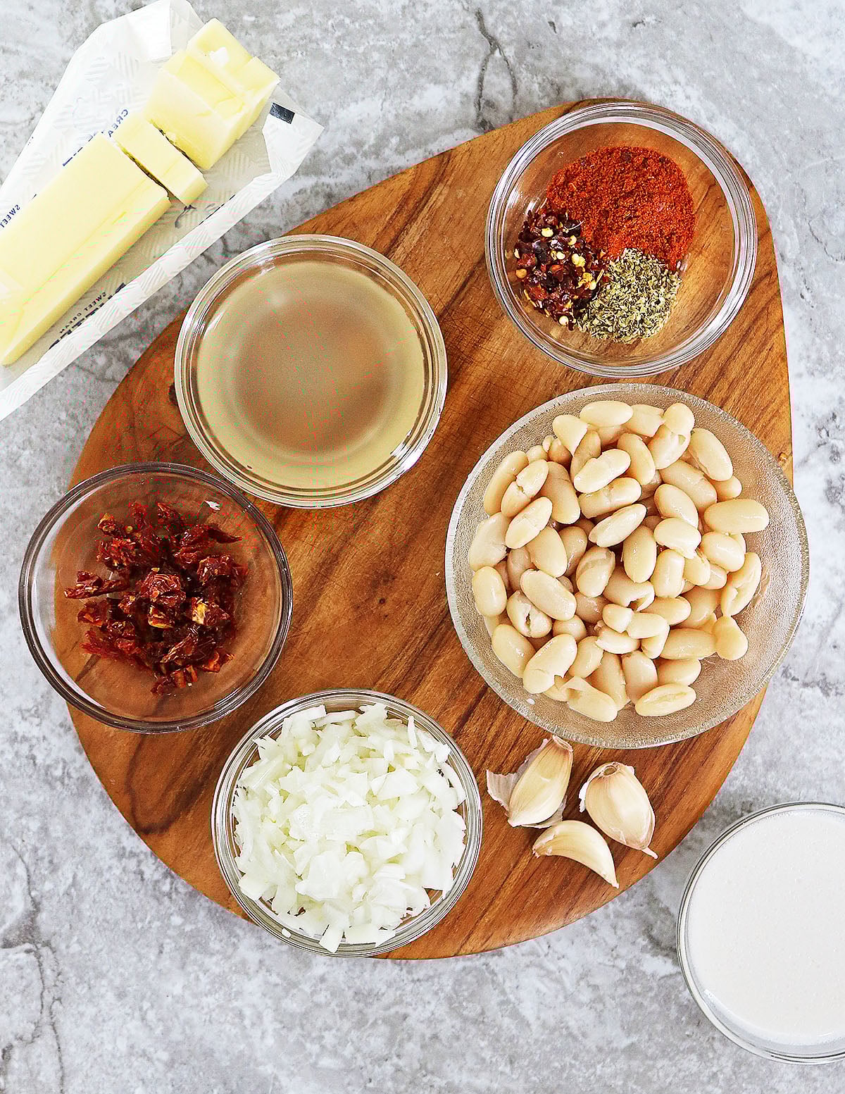 The 10 ingredients to make 15 minute White Bean Marry Me Skillet laid on a wood cutting board on a gray marble counter.