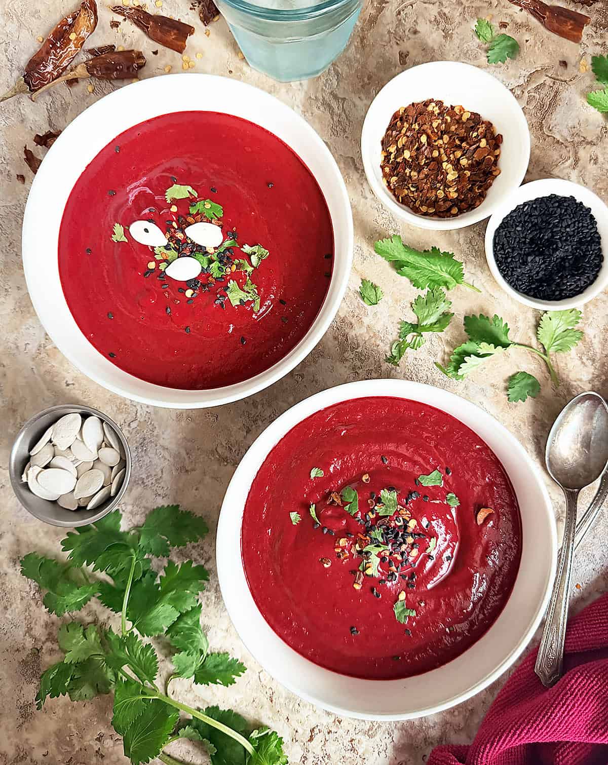 Two bowls of Roasted Beetroot Soup (Creamy, Vegan & Spiced) with pasrley and chili flakes around them.