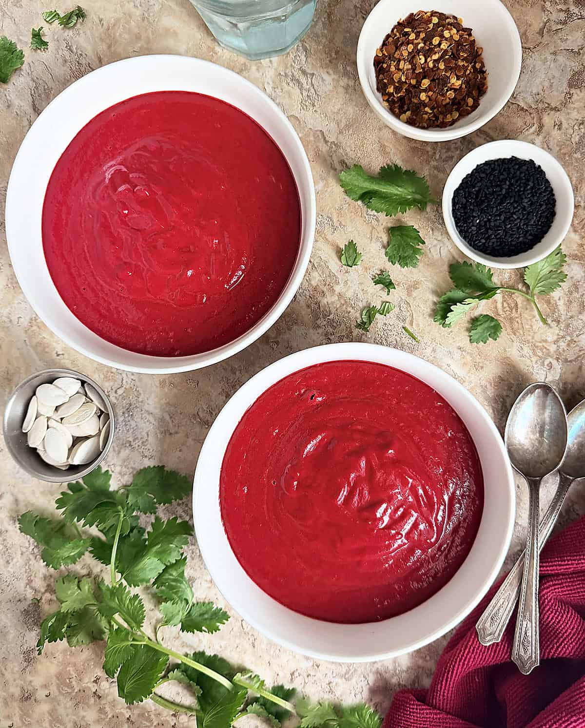 Simple vibrant roasted beet soup in two bowls with fresh cilantro and red pepper flakes around it.